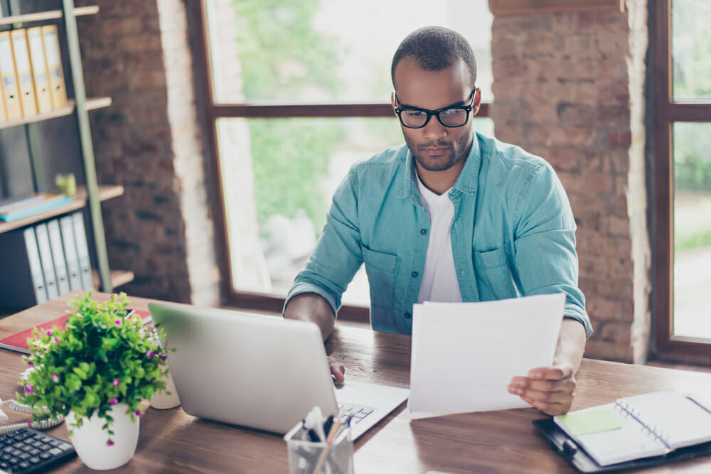 Man sitting at a desk looking at a piece of paper and his laptop