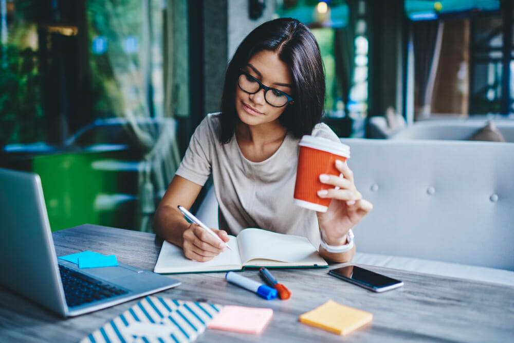 Woman sitting at a table with a notebook, laptop and cup of coffee