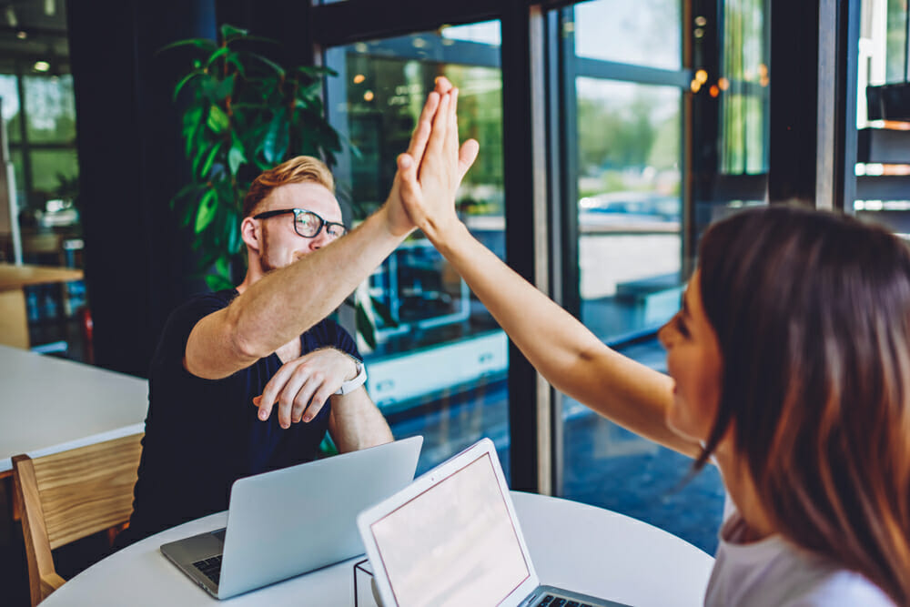 Man and woman sitting at a table with their laptops high-fiving