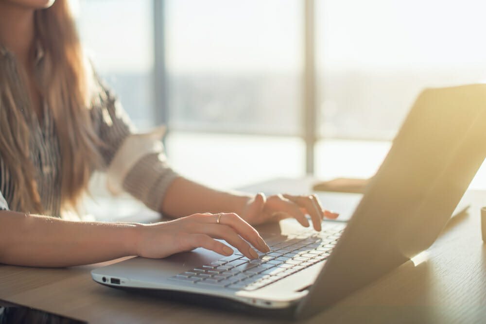 Woman typing on a laptop with a window next to her